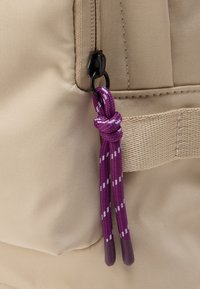 Μπεζ υφασμάτινη τσάντα με μαύρο τράβηγμα φερμουάρ προσαρτημένο σε μοβ πλεγμένο κορδόνι με λευκές ρίγες, δεμένο στον κόμπο στο τέλος.