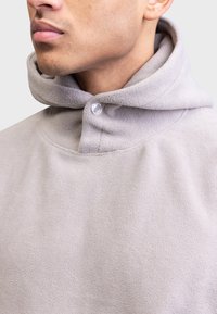 Nahaufnahme einer Person, die einen hellbeigen Fleece-Hoodie mit einem Druckknopf im Nackenbereich trägt.