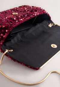 Petit sac pochette ouvert avec sequins rouges, intérieur en tissu noir, fermeture magnétique dorée et chaîne dorée sur surface blanche.