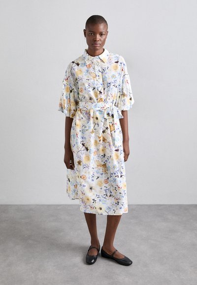 Robe à imprimé floral bleu pâle, avec des manches courtes bouffantes, un col, et une ceinture à nouer à la taille. Portée avec des chaussures plates noires.