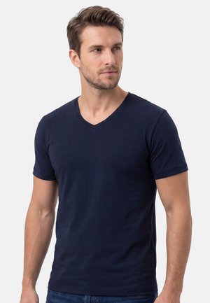 Junger Mann mit kurzen braunen Haaren, der ein schlichtes dunkelblaues V-Ausschnitt-T-Shirt trägt, schaut leicht nach rechts vor neutralem Hintergrund.