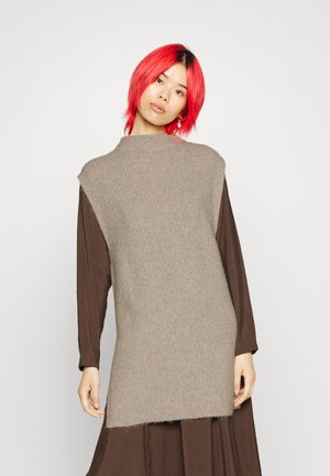 Gebreide mouwloze tuniek in beige met een hoge halslijn en ribbeltextuur, gelaagd over een bruine jurk met lange mouwen.
