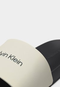 Κοντινό πλάνο ενός μαύρου και λευκού σαγιονάρου Calvin Klein με το όνομα της μάρκας στο λευκό λουράκι.