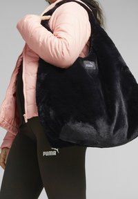 Person wearing a pink jacket and black Puma leggings, holding a large black plush Puma tote bag over their shoulder.