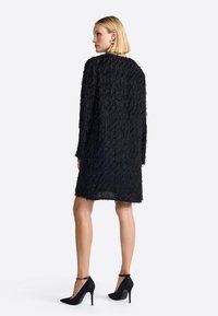 Fekete ruha textúrált rojt dizájnnal, hosszú ujjakkal és laza szabással, fekete magassarkú cipővel párosítva. Simaságú anyag, térd alá érő fazon.