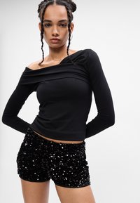 Mujer con el cabello trenzado que lleva una blusa negra de manga larga y hombros al descubierto, y pantalones cortos negros con lentejuelas, de pie frente a un fondo claro y liso.