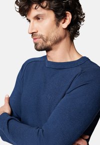 Granatowy sweter dzianinowy z teksturowanym ściągaczem, okrągłym dekoltem i małym haftowanym logo na piersi. Długie rękawy z dopasowanymi mankietami.