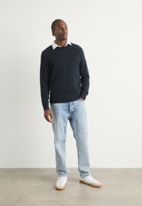 Maglione di lana blu navy sopra una camicia bianca con colletto, abbinato a un paio di jeans in denim azzurro e sneakers bianche con suole marroni, in piedi contro uno sfondo neutro.