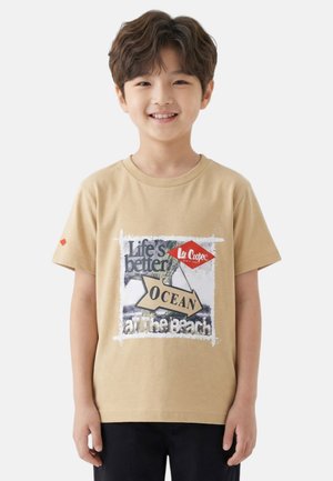 Garçon souriant aux cheveux bruns ondulés portant un t-shirt beige avec l'inscription « La vie est meilleure OCEAN à la plage » et un pantalon noir, debout devant un fond blanc.