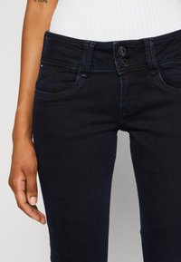 Tmavě modré džíny s přiléhavým střihem, obsahující dvě přední kapsy, zapínání na knoflík a prošití podél pasu a kapes.