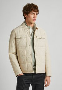 Beige quilted jacket with a collared neckline, snap buttons, and two front pockets, layered over a light grey zip-up top.