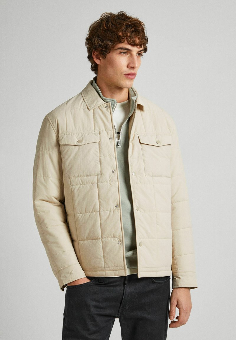 Beige quilted jacket with a collared neckline, snap buttons, and two front pockets, layered over a light grey zip-up top.