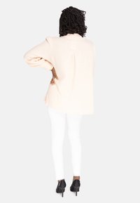 Camicia oversize di un rosa pallido con maniche lunghe, tessuto strutturato, dettaglio pieghe sul retro, indossata sopra pantaloni skinny bianchi, abbinata a tacchi neri.
