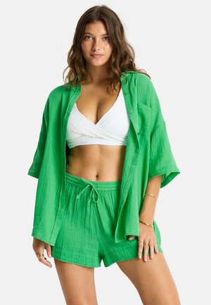 Femme aux cheveux bruns ondulés portant une chemise verte ample boutonnée, un short vert assorti et un crop top blanc noué, debout sur fond blanc.