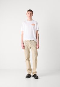 T-shirt bianca collo tondo con logo arancione, abbinata a jeans beige chiaro e calzature nere. Modello in piedi contro uno sfondo neutro.