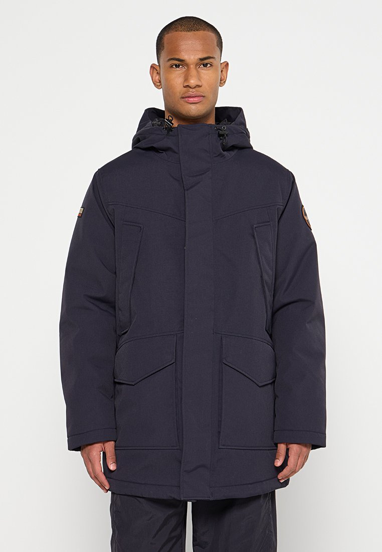 Man die een donkerblauwe winterparka met capuchon en meerdere voorkruizen draagt, staand tegen een effen witte achtergrond.