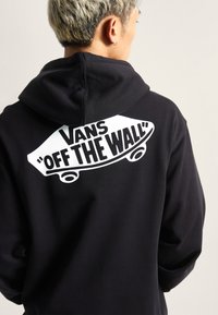 Felpa nera con cappuccio e grande grafica bianca di uno skateboard con il testo "VANS 'OFF THE WALL'" sul retro. Tessuto liscio, cappuccio con lacci, vestibilità comoda.