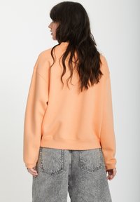 Sweatshirt léger orange avec une coupe décontractée et un ourlet côtelé, associé à un pantalon en denim gris doté de poches arrière et d'une texture délavée.