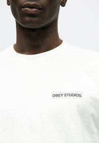 Κοντινό πλάνο ενός ατόμου που φοράει λευκό πουκάμισο με την επιγραφή "OBEY STUDIOS" σε μαύρο στο στήθος.