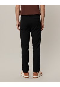 Schwarze Hosen mit maßgeschneidertem Schnitt, zwei Taschen hinten und einer glatten Textur. Kombiniert mit braunen Sneakers und einem braunen Oberteil.