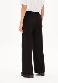 Pantalons larges noirs avec une taille élastique, fabriqués dans un tissu lisse. L'ourlet présente une finition soignée, assortie à de grandes chaussures noires.
