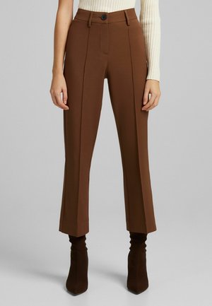 Femme portant un pantalon marron taille haute ajusté, coupé court avec des coutures sur le devant et des bottines marron à talons sur un fond gris.