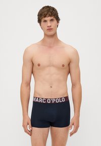 Férfi sötétkék fürdőnadrág bordó rugalmas derekú, fehér "MARC O’POLO" felirattal. Simaságával és testhezálló kialakításával közepes magasságú.