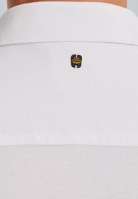 Weißes Poloshirt mit einem schwarzen und goldenen Logo-Tag auf der Rückseite. Der Stoff ist glatt und hat ein klassisches Kragen-Design.