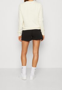 Person som bär en krämfärgad långärmad tröja, svarta shorts, vita Nike-strumpor och vita sneakers, stående vänd mot en enfärgad vägg.