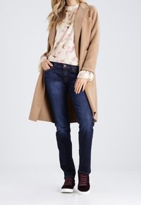 Beige ullblandad kappa över en blomsterprydd krämfärgad topp, mörkblå jeans och vinröda sneakers med vita sulor. Långa ärmar och figursydd design.