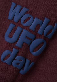 Bordowy materiał z wyraźnym, wypukłym niebiesko-granatowym napisem „Światowy Dzień UFO”. Tekstura jest dziana, z widocznymi detalami ściegów.