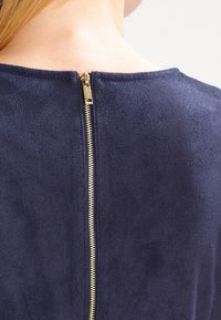 Top en daim bleu marine avec une fermeture éclair dorée apparente à l'arrière, une texture lisse et un col arrondi.