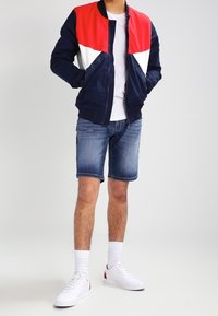 Cazadora bomber marino con detalles en rojo y blanco, combinada con pantalones cortos de mezclilla. Las zapatillas blancas y los calcetines hasta el tobillo completan el look.