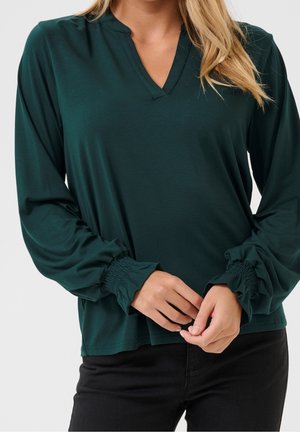 Femme portant un chemisier vert foncé à manches longues avec des poignets froncés et un pantalon noir, mains jointes devant.