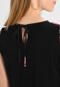 Vue arrière d'une femme aux cheveux châtain clair portant un haut noir avec une ouverture nouée dans le dos et un bord floral aux manches.