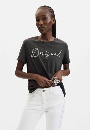 Donna con i capelli intrecciati che indossa una t-shirt nera con il testo "Desigual" e pantaloni bianchi, si sistema la camicia, indossa un grande anello decorativo.