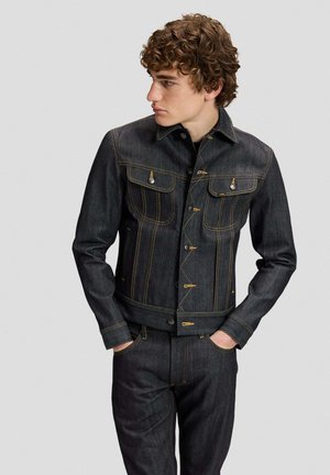 Junger Mann mit lockigem Haar, der eine dunkle Jeansjacke und passende Jeans trägt, Hände in den Taschen, blickt nach links vor schlichtem Hintergrund.