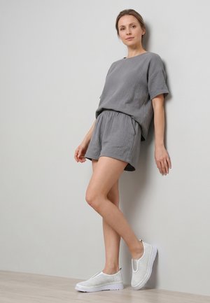 Kvinde læner sig mod en væg iført løs grå kortærmet top og shorts med hvide slip-on sneakers mod en ensfarvet baggrund.
