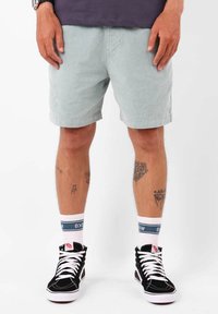 Shorts en velours côtelé vert clair, associés à des baskets noires et des chaussettes blanches avec des accents bleus. Tatouages visibles sur les jambes.