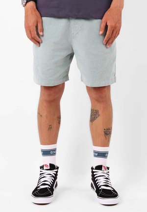 Shorts en velours côtelé vert clair, associés à des baskets noires et des chaussettes blanches avec des accents bleus. Tatouages visibles sur les jambes.