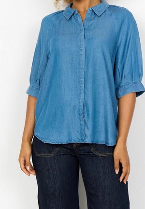 Femme portant une chemise bleue ample boutonnée avec des manches jusqu'au coude et un jean en denim bleu foncé sur un fond uni.