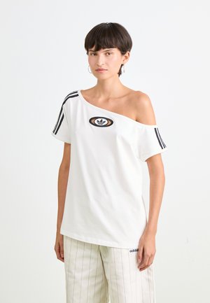 Moteris dėvi baltą Adidas marškinėlį be pečių su logotipu ir juodomis juostelėmis, derindama su baltais juostuotais Adidas kelnėmis prieš neutralų foną.