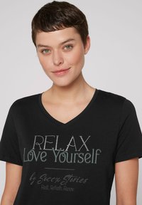 Camiseta negra de cuello en V con el texto "RELÁJATE, ámate a ti mismo" y acentos decorativos. Fabricada con tela suave y un ajuste relajado.