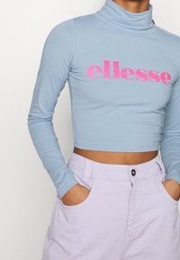 Himmelblaues, langärmliges Rollkragenoberteil mit einem pinken "ellesse"-Logo. Kombiniert mit hochgeschnittenen lavendelfarbenen Shorts mit frontalen Taschen.