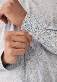 Hvid skjorte med et blåt, gult og grønt blomsterprint. Manchetknappen er rullet op, så hvide knapper og struktureret stof ses.