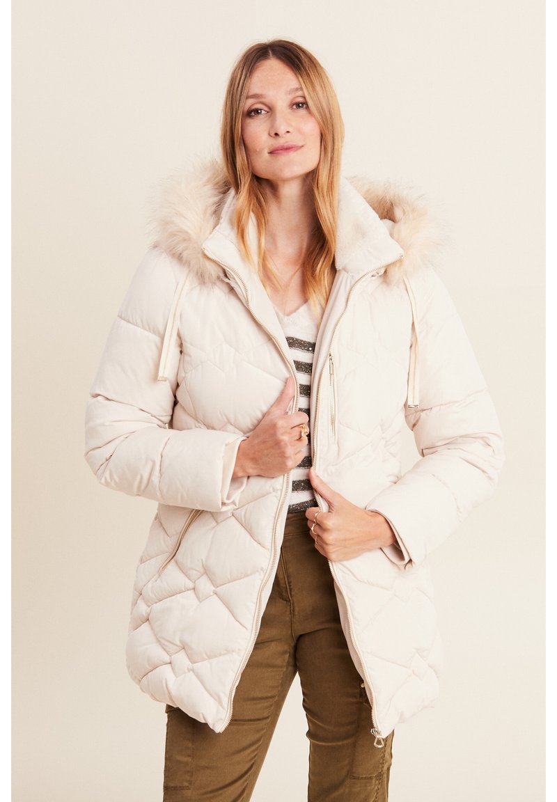 Giacca bianca voluminosa con cappuccio foderato in finta pelliccia, motivo a rombi, zip frontale e tasche laterali. Modella con pantaloni verde oliva.