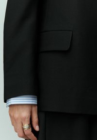 Veste de costume noire avec une poche frontale, associée à une chemise à manches avec des rayures bleues et blanches, main ornée d'une bague en argent.