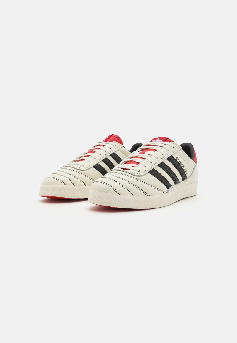 Sneaker in pelle bianca con strisce nere, dettagli rossi e superficie texture. Punta arrotondata e suola in gomma. Presenta lacci e dettagli con logo.