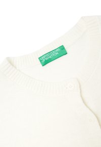 Bílý pletený cardigan s kulatým výstřihem, žebrovaným lemem a dvěma knoflíky. Štítek nese nápis "United Colors of Benetton" v zelené barvě.