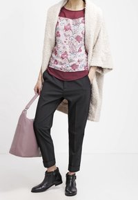 Cardigan beige doux porté sur un haut à motif floral rose et bleu, associé à un pantalon noir fuselé et des bottines noires, tenant un sac rose.